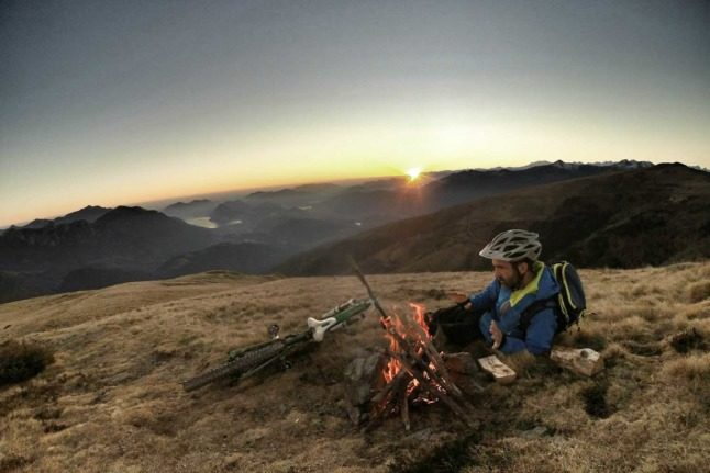 A man sits with a campfire and his bike in the mountains in Ticino, Switzerland. Photo by Graziano De Maio on Unsplash