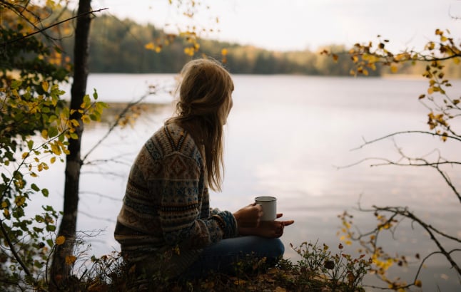 A woman sitting by a lake, holding a cup of warm beverage. It's autumn in Sweden.