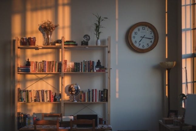 A half-lit bookshelf with books and a vase of flowers next to a wall clock