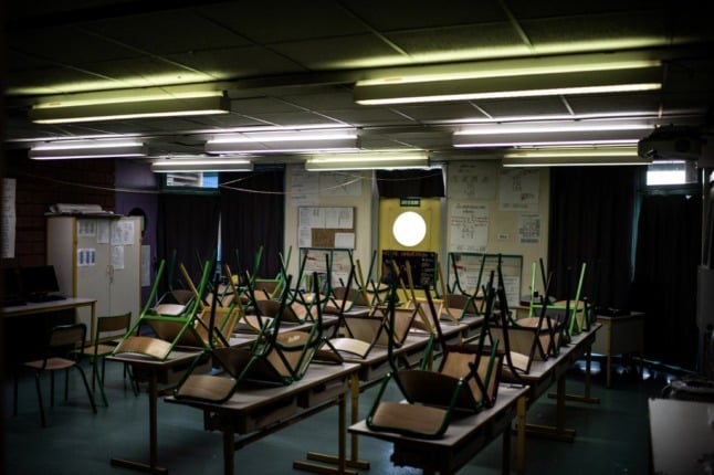 An empty school classroom, with chairs stacked on desks and overhead electric lights switched on