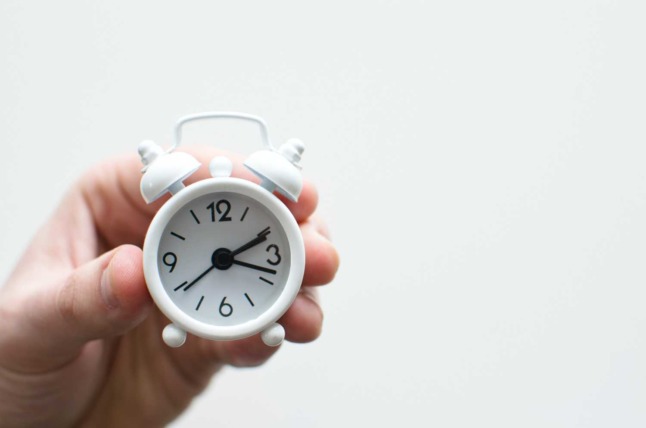 A person holds an unusually small white clock against a blank background. 