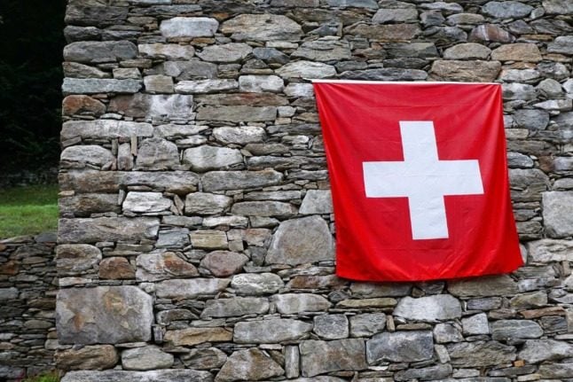 A Swiss flag against a stone house somewhere in the Swiss mountains