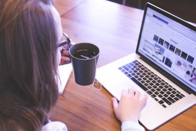 A woman drinks a cup of tea while looking at her laptop