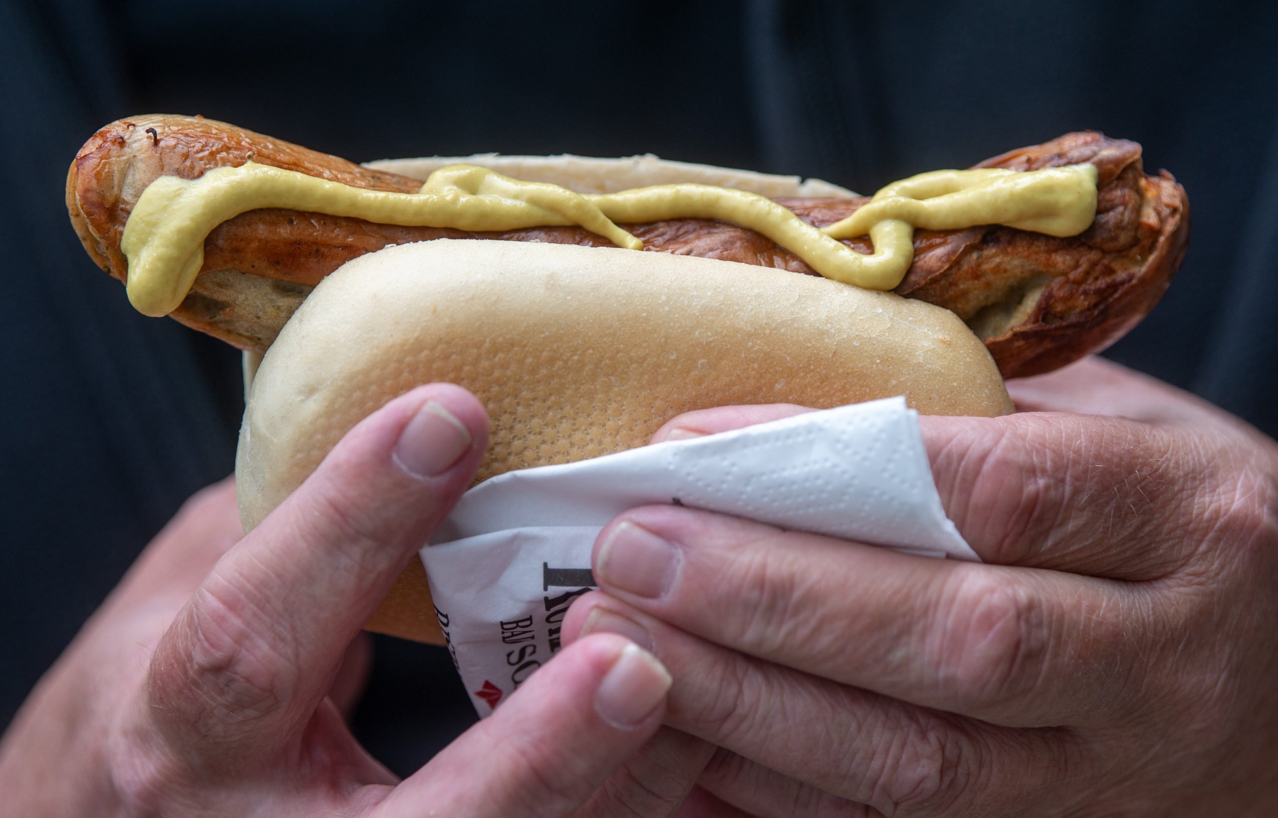 A man holds a bratwurst covered in mustard.