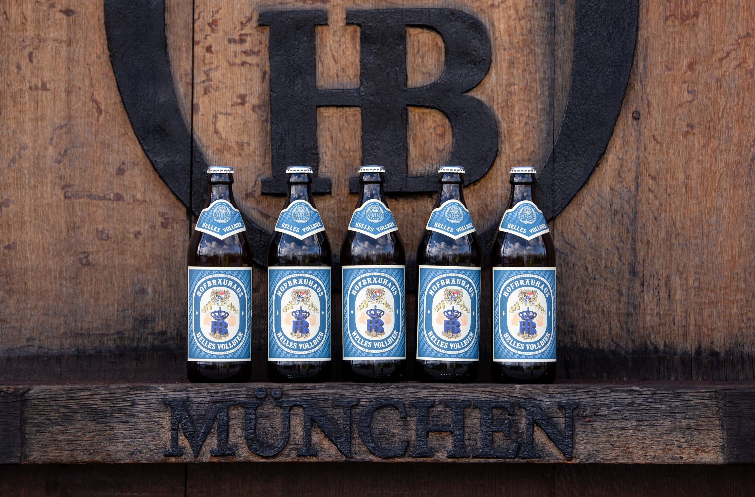 Beer bottles from the Hofbräuhaus Munich stand in front of a logo of the brewery on the brewery's premises.