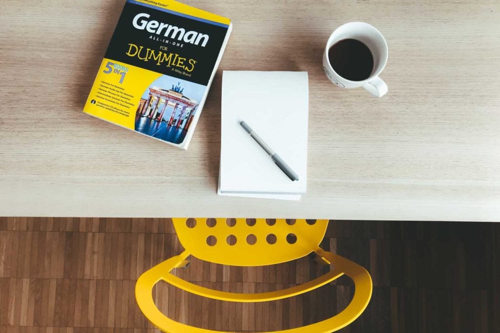 A German for Dummies language book sits atop a desk next to a pen and a cup of coffee. Photo by Jan Antonin Kolar on Unsplash