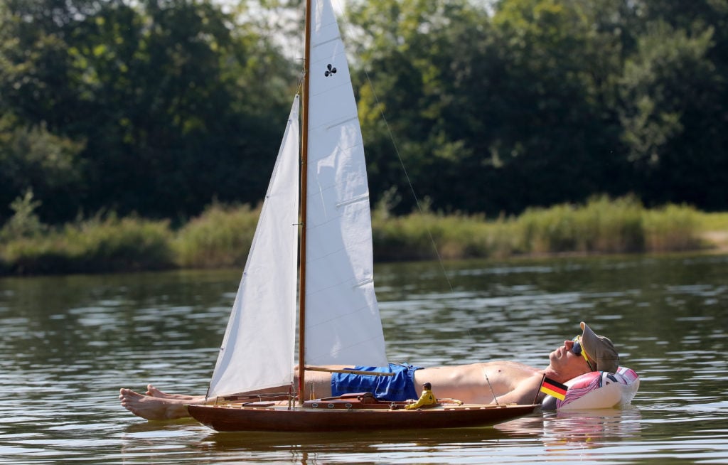 A man on an air mattress sunbathing on a lake while a model boat passes him by.