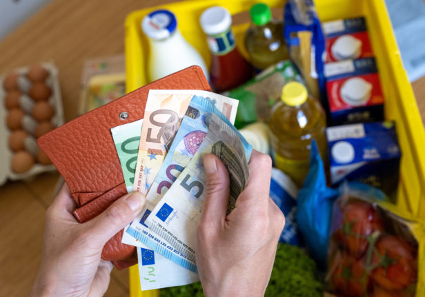 A box of groceries sits on a kitchen table while a woman holds euro banknotes in her hands.