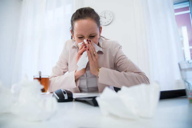 A woman blows her nose at her desk in the office.