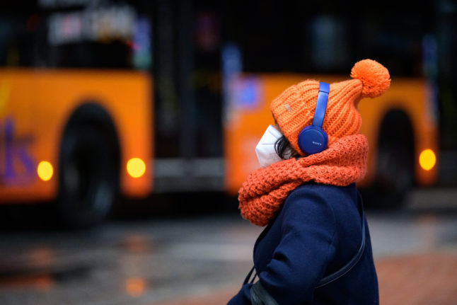 A woman wearing an FFP2 mask, thick cap and headphones sits at a bus station as a public bus passes by.