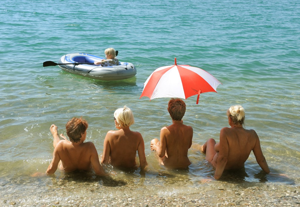 Four nude women sit on the edge of lake and face away under the sun.