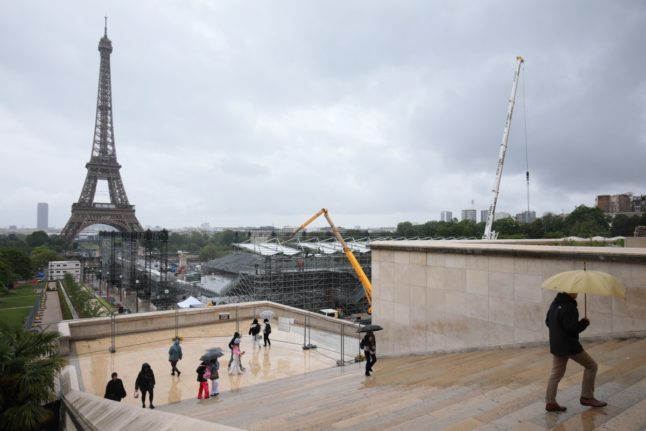 Pedestrians holding umbrellas walk past the temporary venue under construction for the upcoming Paris 2024 Olympic Games, with the Eiffel Tower in the background,