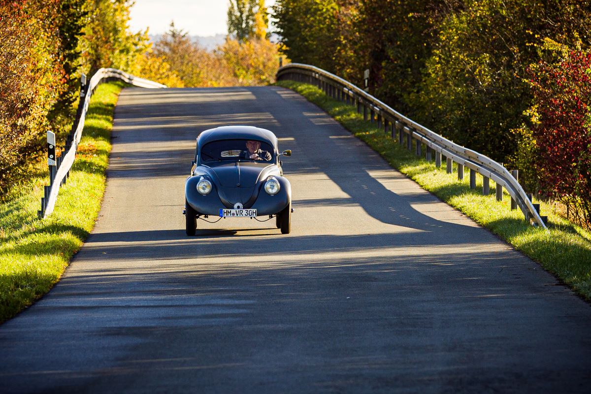 A grey VW Beetle drives in sunshine along a country road