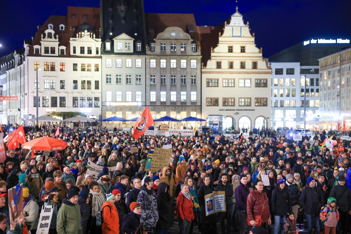 A large crowd is gathered in Leipzig city square at night with protest sings and red flags.