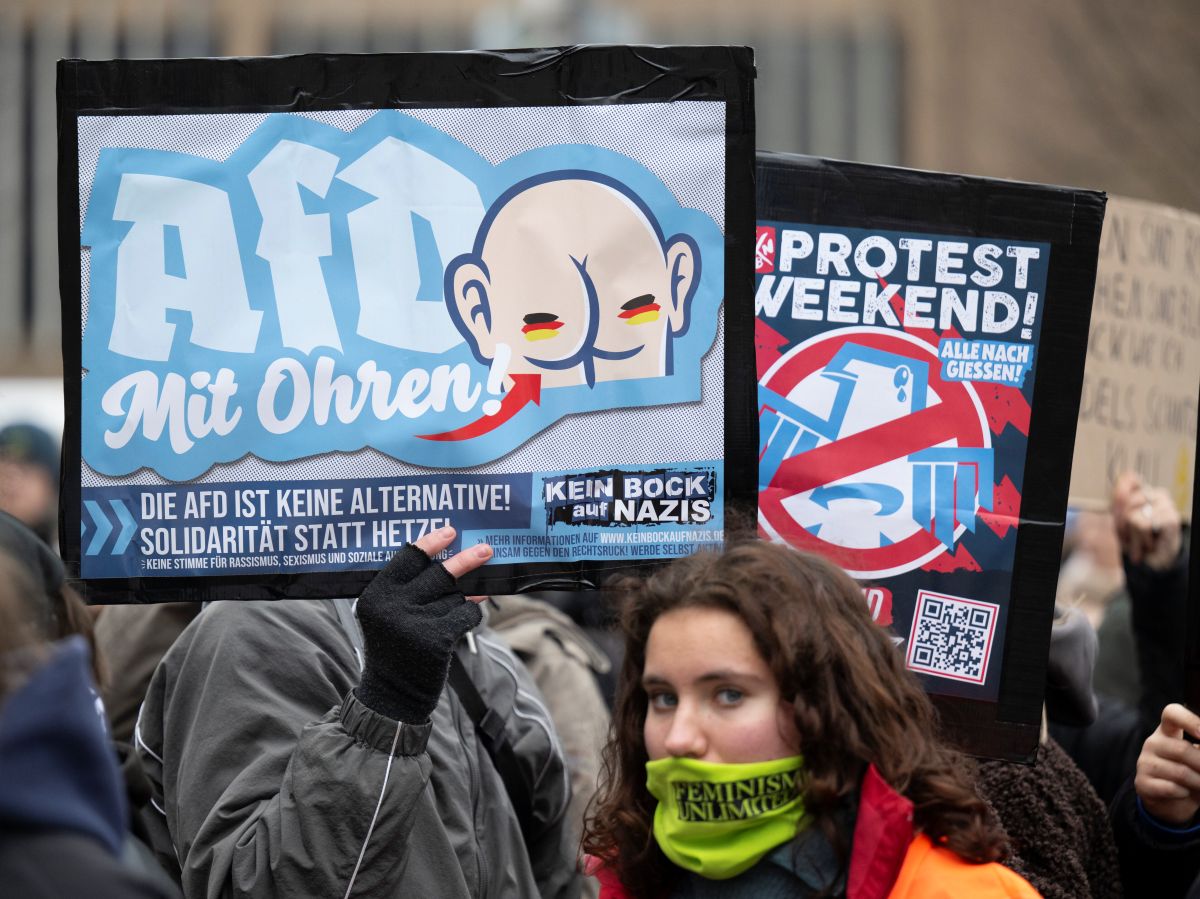 A young woman smiles behind a bandana in front of protest signs against the AfD.
