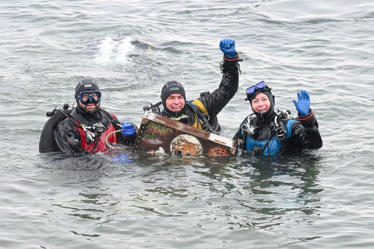 Scuba divers hold up a treasure chest in the water and wave to the camera.