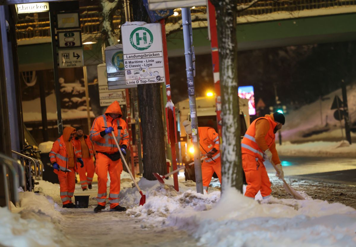Men in orange suits work to shovel snow from a bus stop in the dark early morning hours.