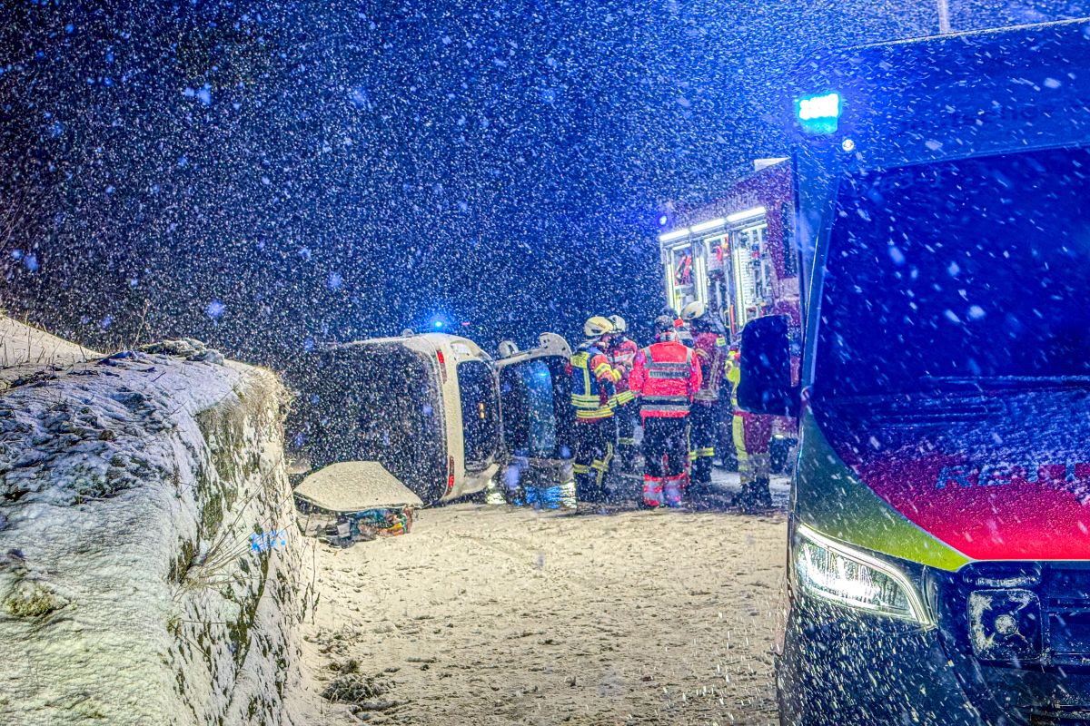 Snow falls heavily on a road at night where emergency workers gather around a car overturned.