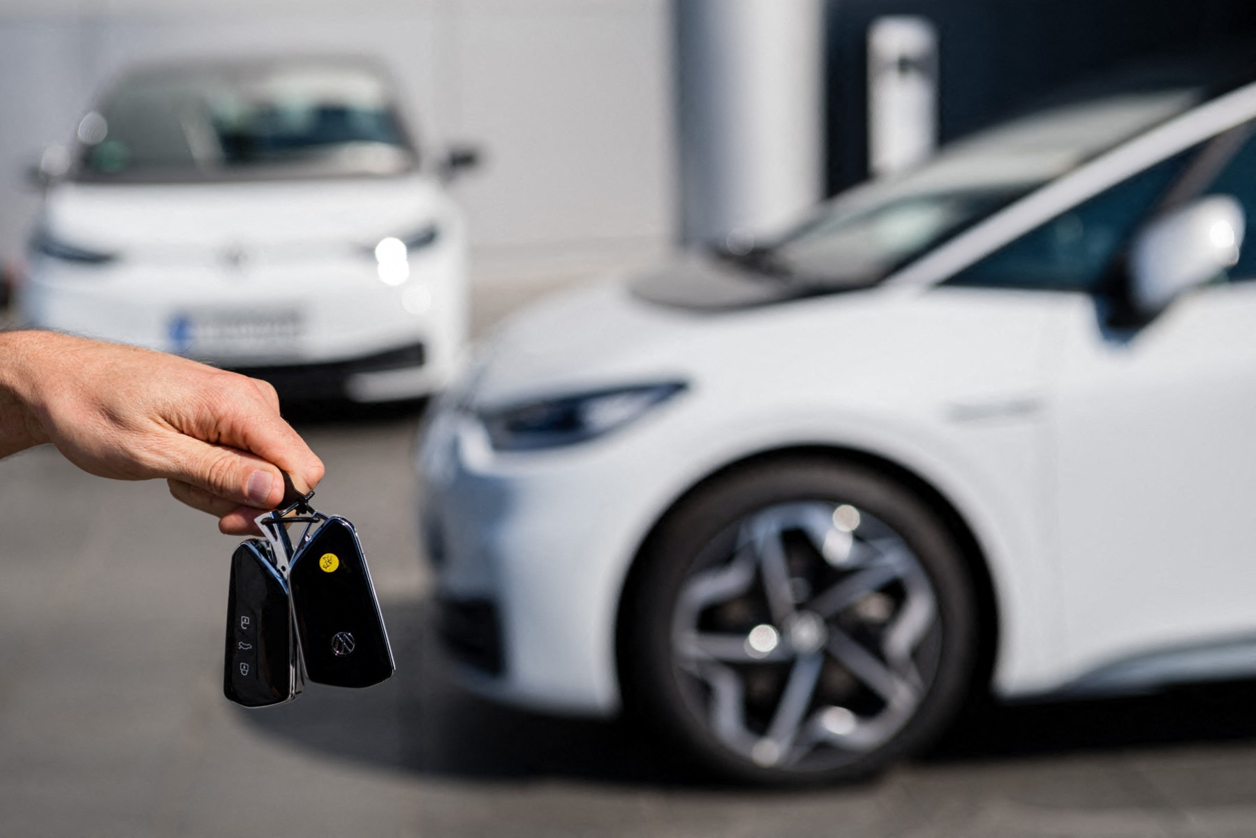 A hand holds a pair of new car keys with a couple new VW Evs parked in the background.