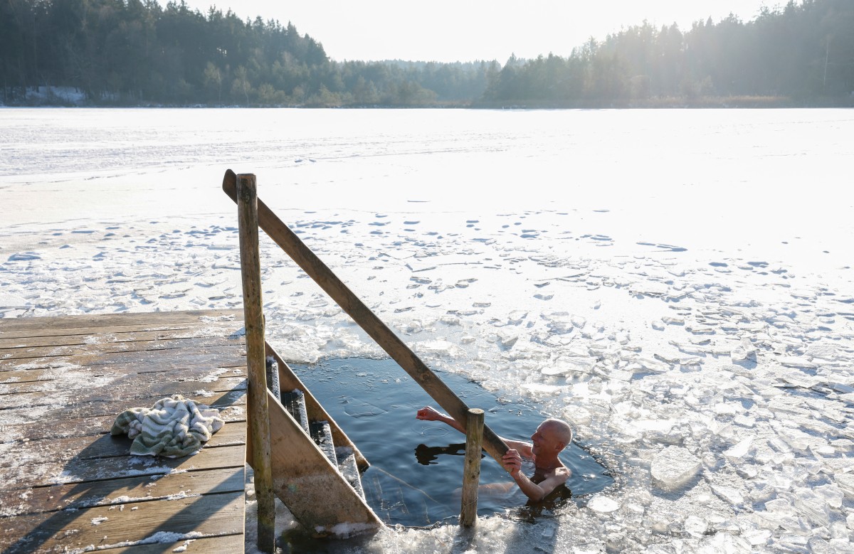A man's head is above the water in an icy lake, he holds onto the handrail of some steps entering the water.