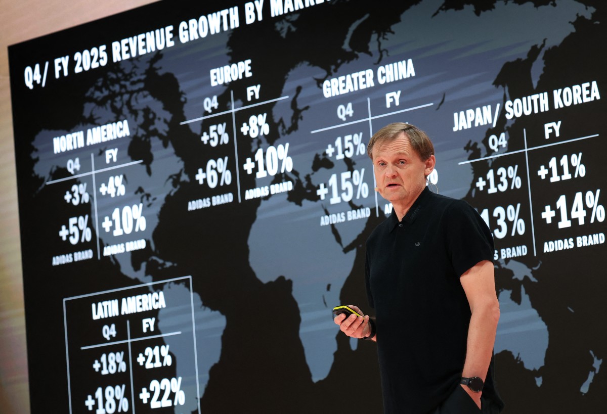 A man holds a power point clicker and stands in front of a world map with percentages representing profit margin increases laid over various regions.