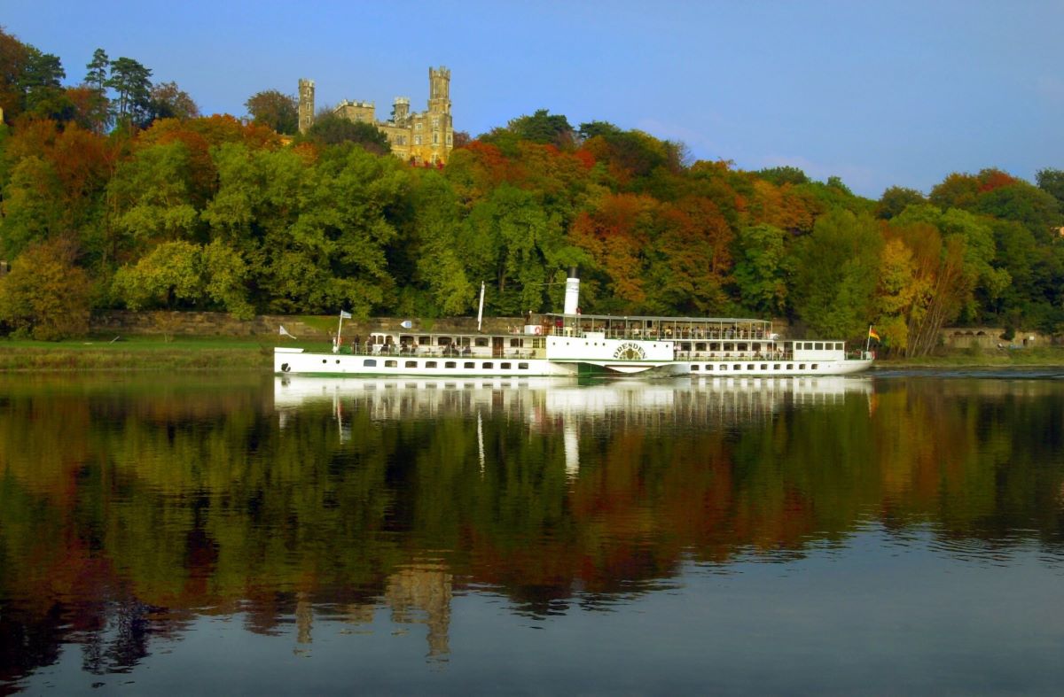 Under a bright blue sky, the passenger steamboat “Dresden” sails past the autumn-colored trees on the banks of the Elbe and Eckberg Castle near Dresden 