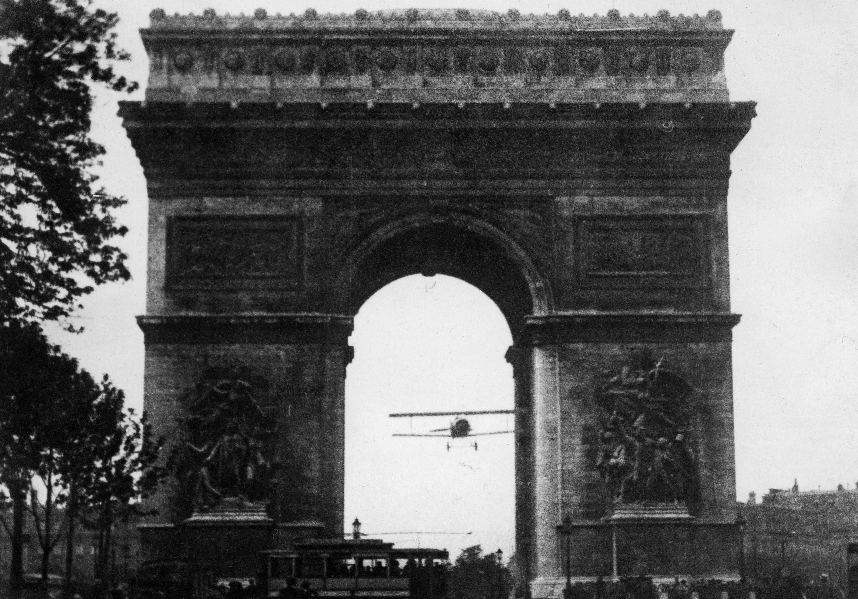Black and white photo of a biplane flying through the Arc de Triomphe arch, the clearance space is very small