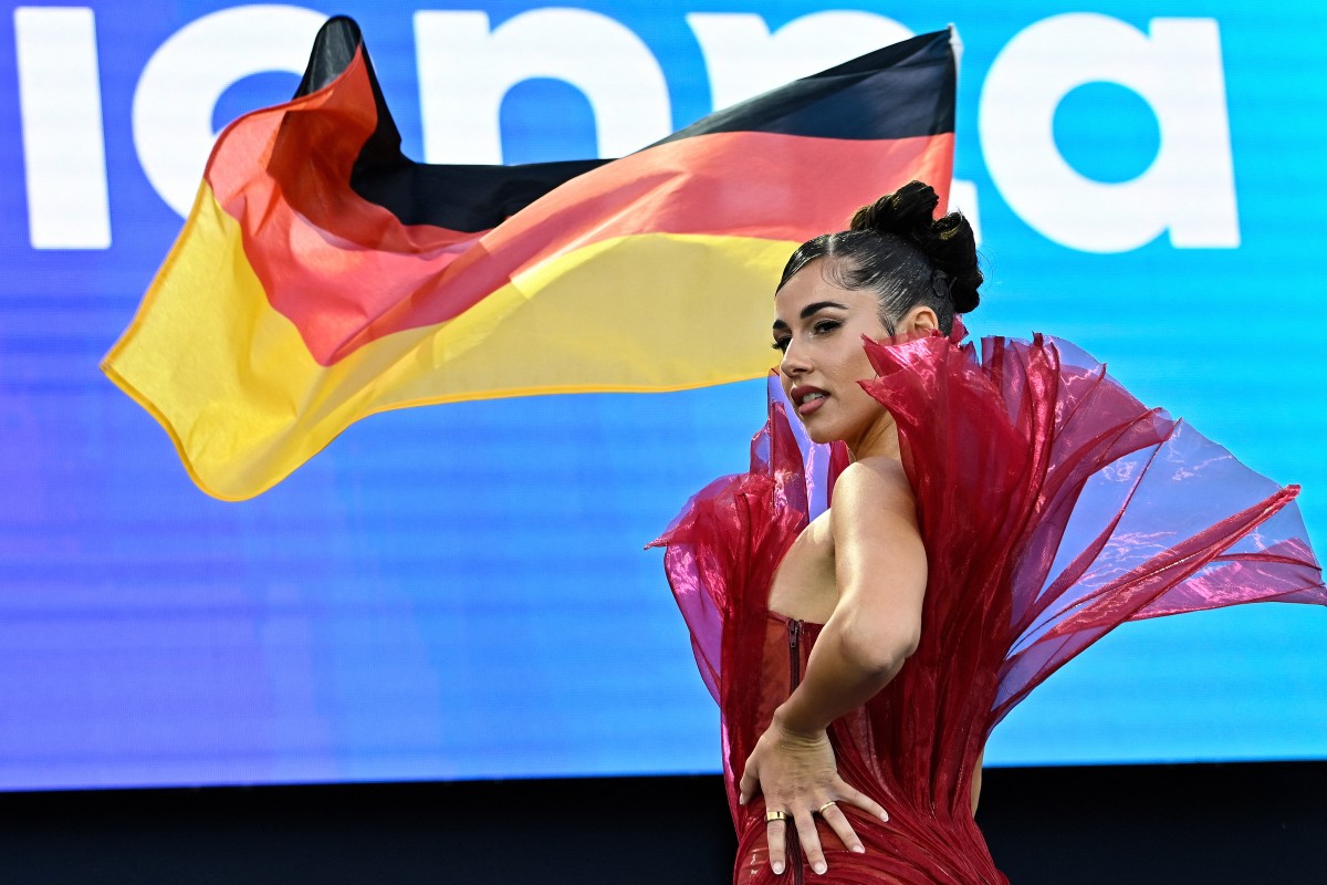 A woman in a red dress stands before a blue backdrop holding the German flag.