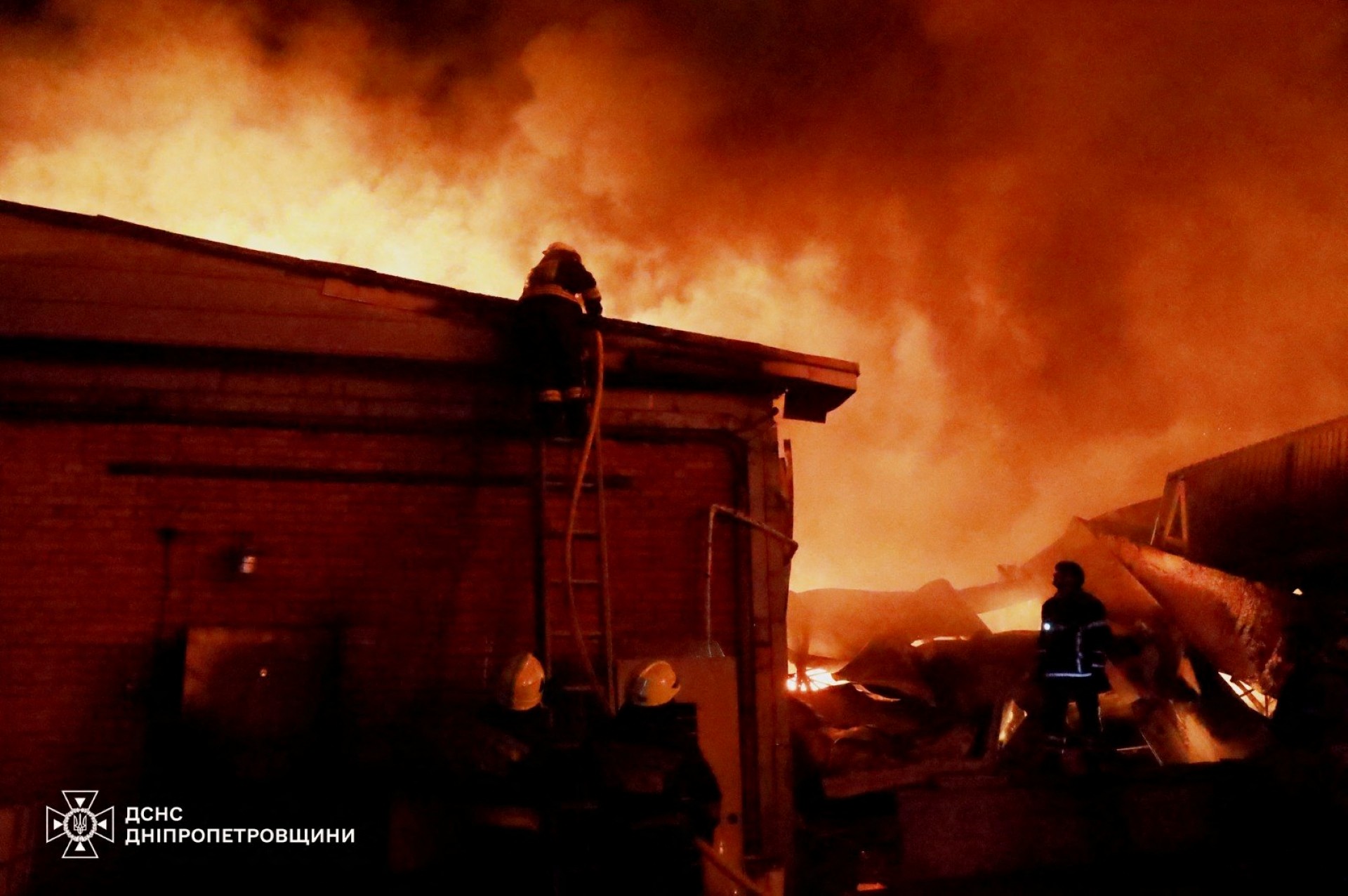 Firefighters are seen in a red-lit night scene with a hose leading up to a building's roof.