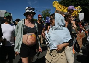 A pregnant demonstrator (2ndL) walks with a painting depicting US President Donald trump holding a bloody axe on her belly during a rally of a "No-G7" coalition