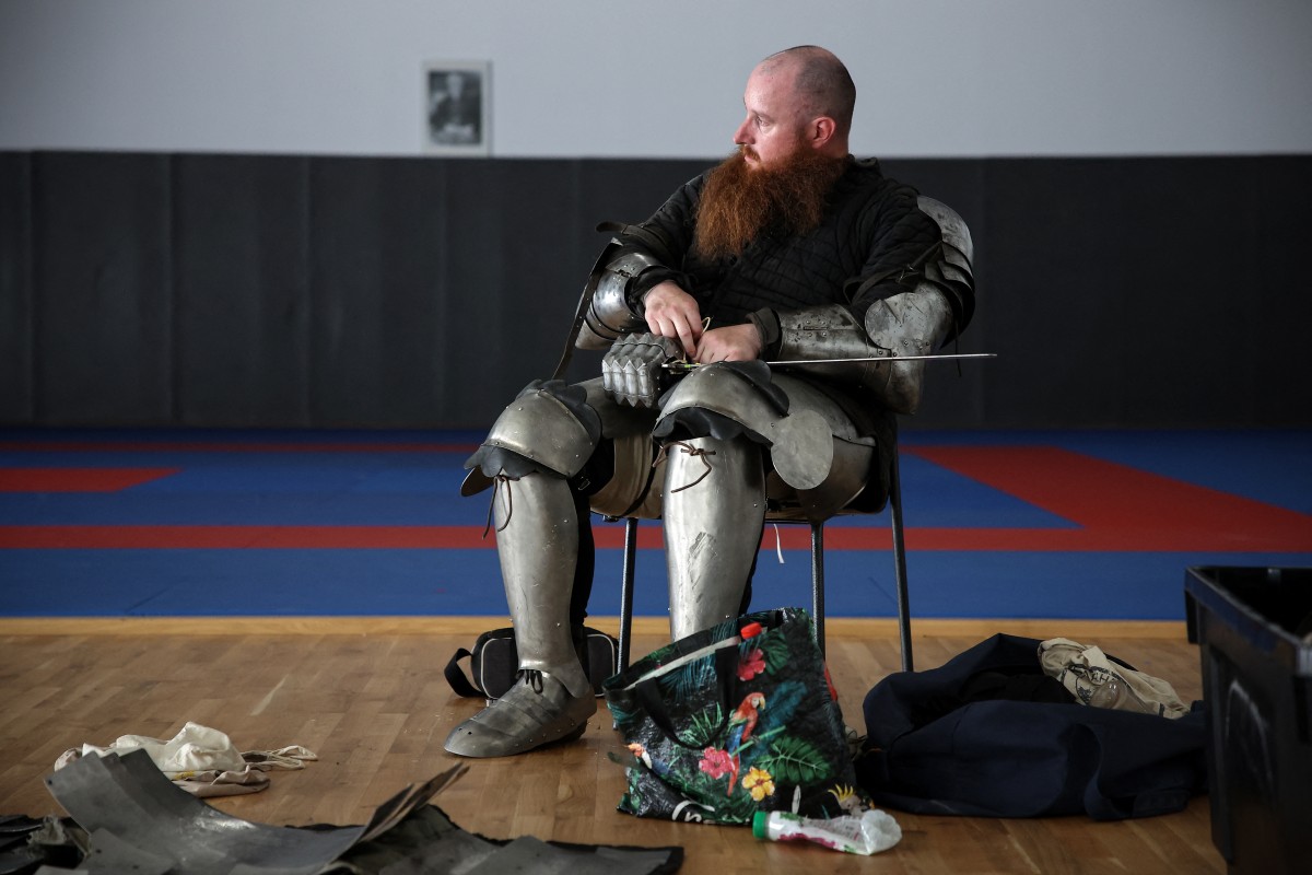 A competitor sits as he waits before fighting. - he wears full armour and holds a sword but his head (shaved with a large beard) is bare. He sits in a room that looks like a school sports hall
