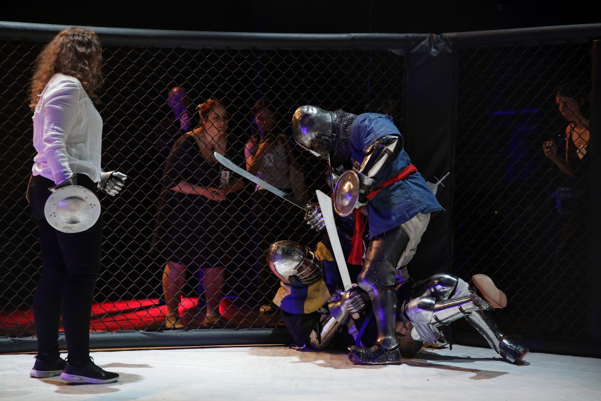 Competitors fight in an MMA (mixed martial arts) arena - one is down on the ground, the other standing over them with a sword