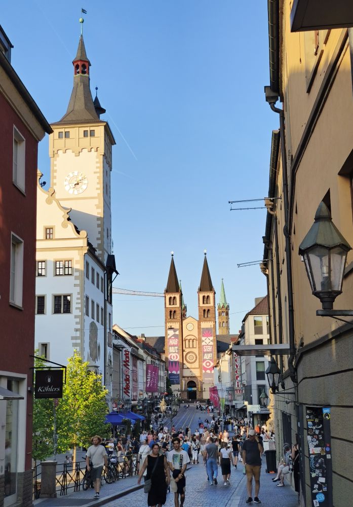 View of a pedestrian street in Würzburg.  People walk in front of a church and tall clock tower.