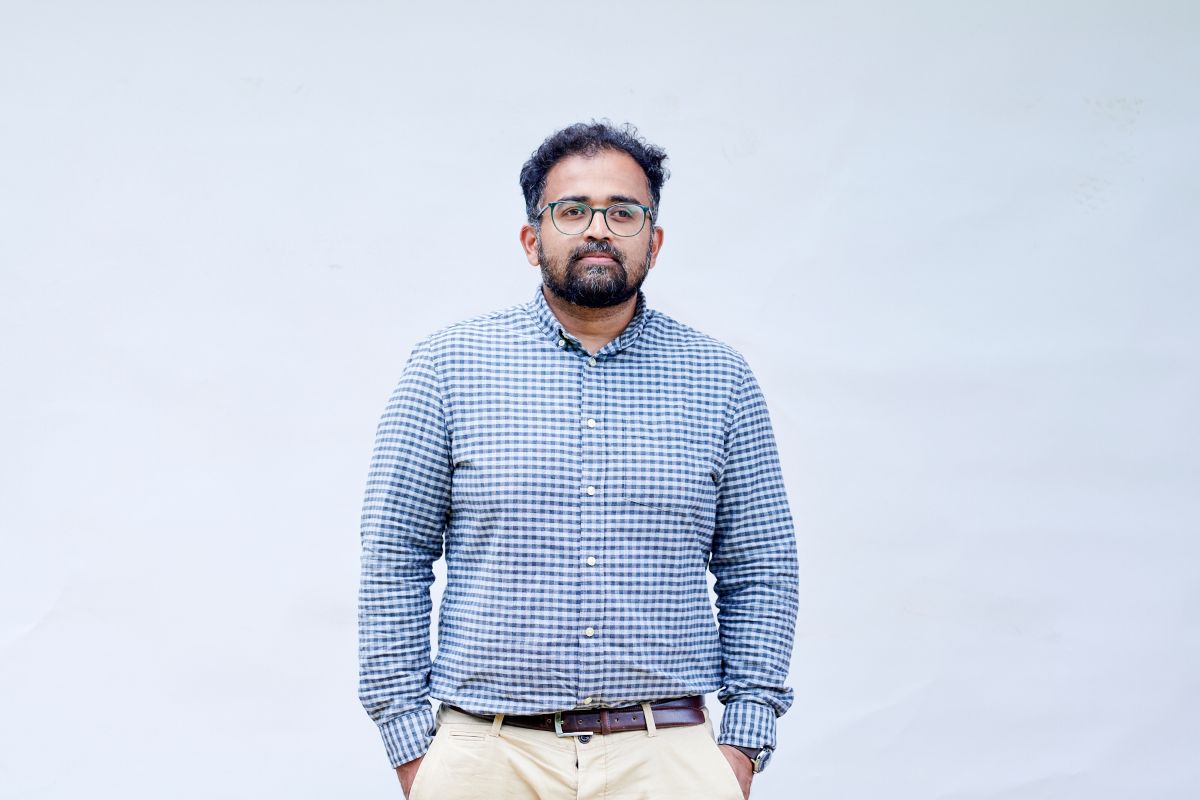 Aju John stands before a white backdrop in a checked button up shirt.
