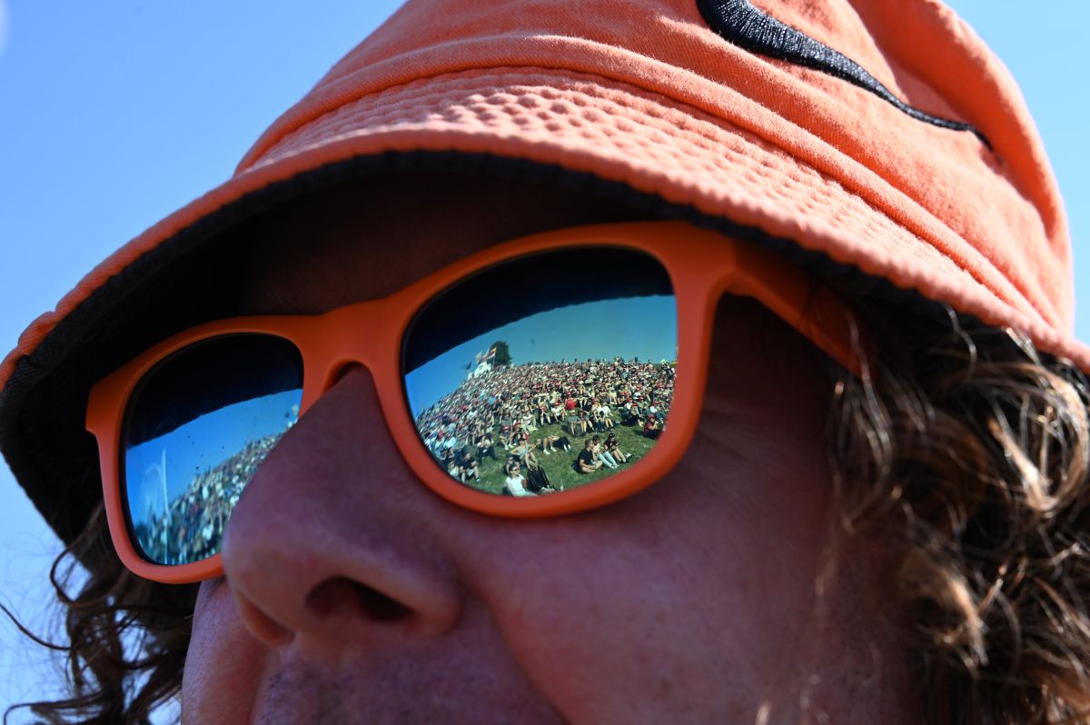 A colorful crowd of people sat on a grassy hill are reflected in a man's orange-rimmed sunglasses.