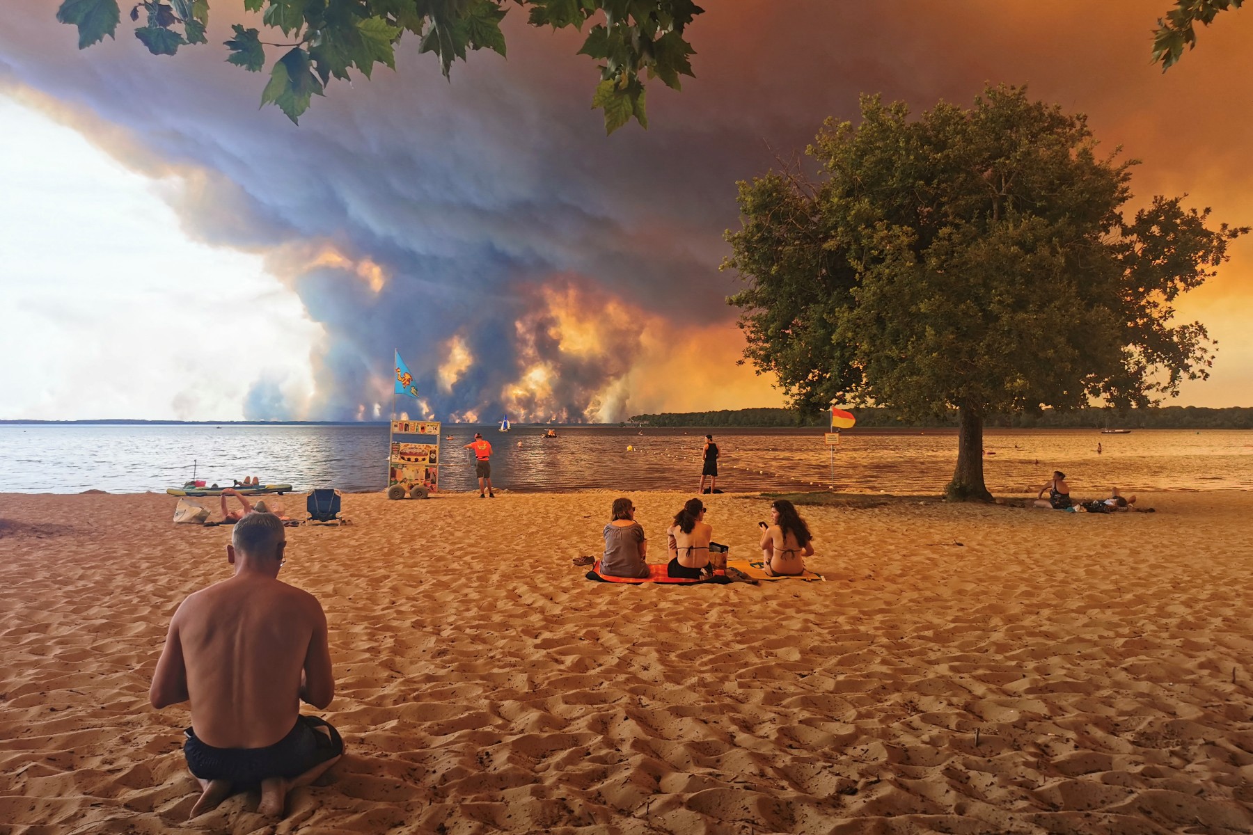 Around a half dozen people scattered, mostly seated, on the beach at the smoke rises in the background