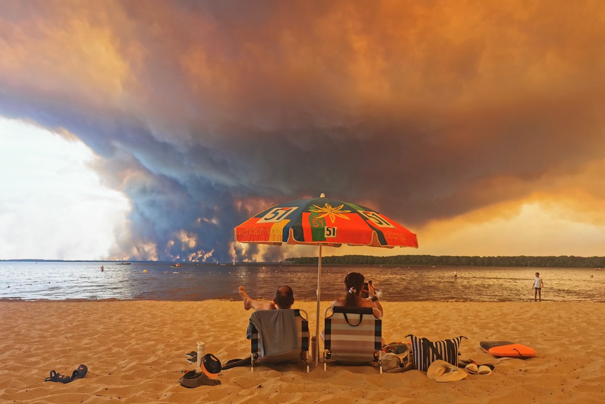 Beach goers sit under a parasol and watch smoke rising from wildfires in the distance