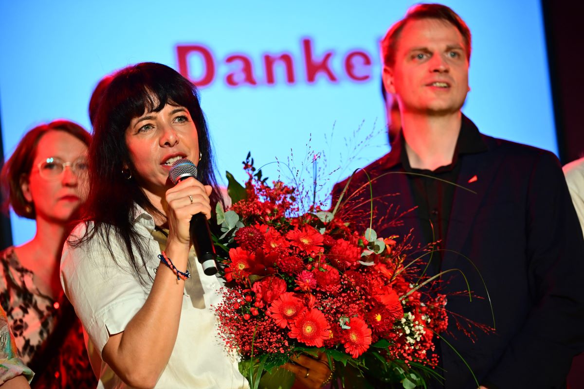 A woman speaks into a micrphone and holds a bouquet of red flowers