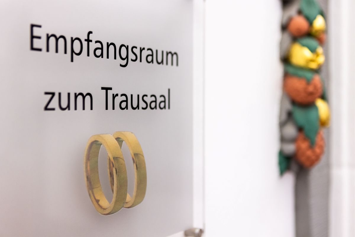 A sign on a door reads 'empfangsraum zum Trausaal' and has a photo of two wedding rings.