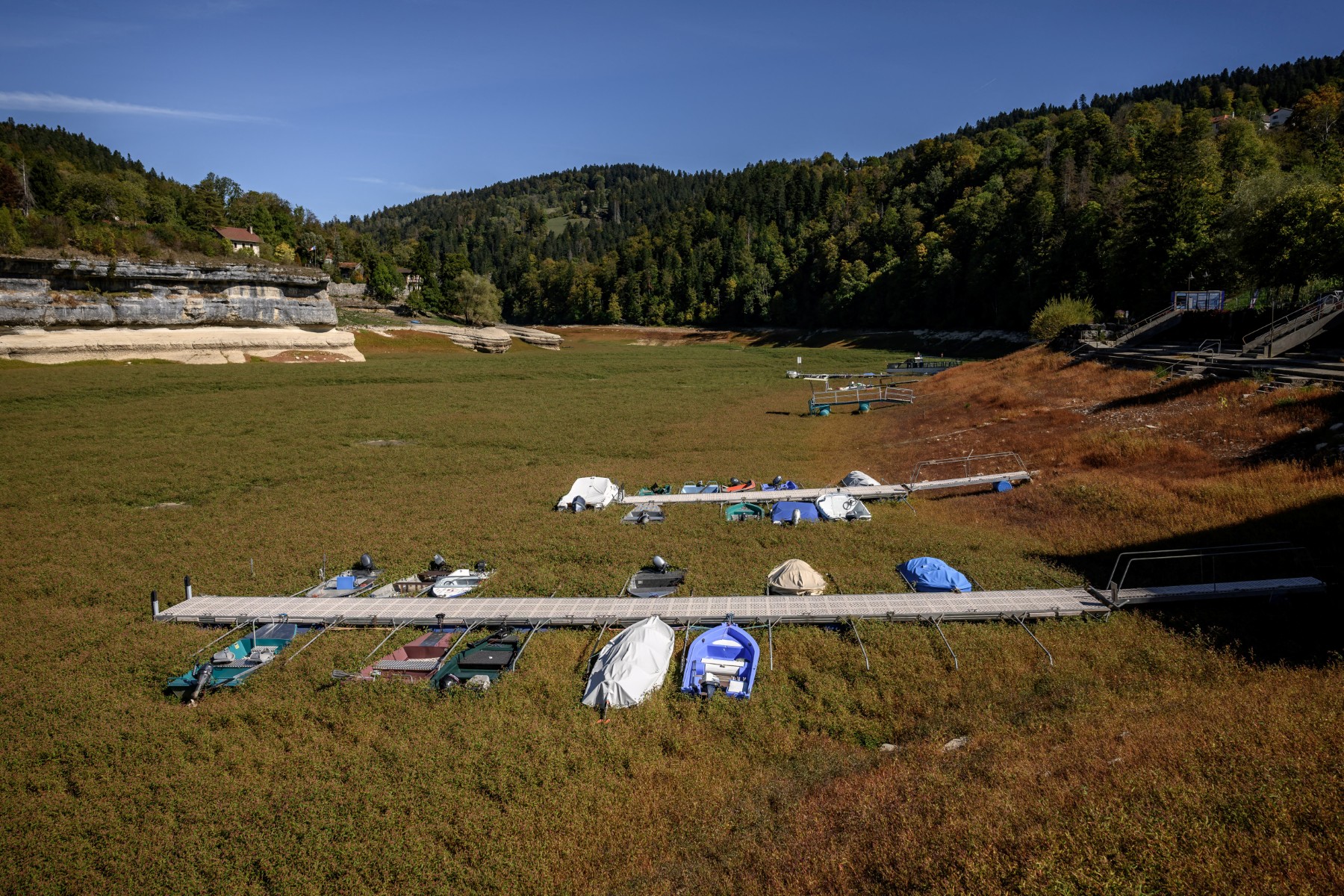 This photograph shows the bed of Lake Brenets (Lac des Brenets), priorly parched and now covered in grass, and a part of the Doubs River forming a natural border between eastern France and western Switzerland, in Les Brenets